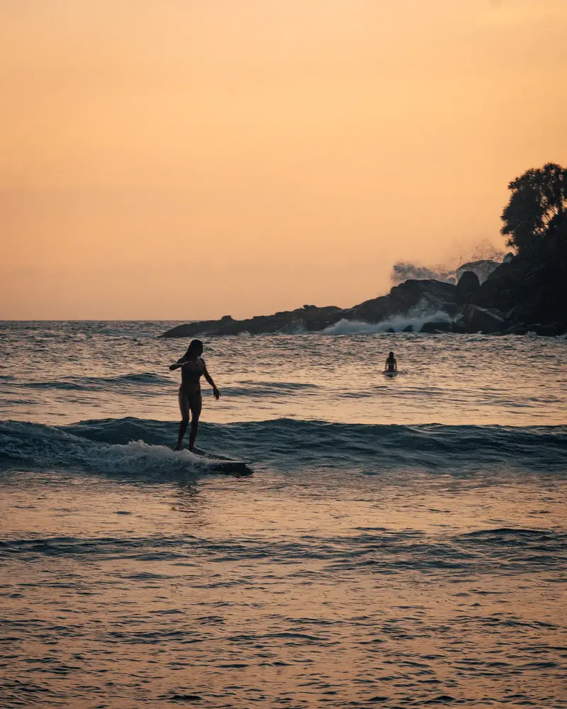A surfer riding a small wave at sunset below a rocky headland