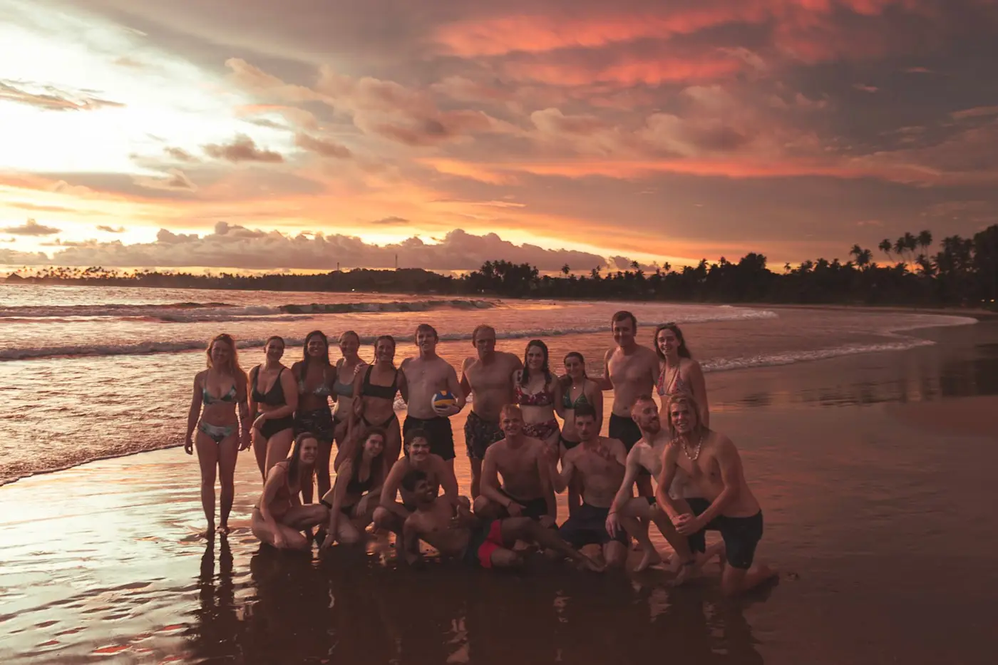 About twenty guests standing together in the shallows at Hiriketiya under a pink sunset sky