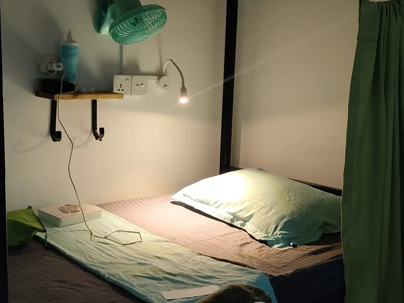 A made-up dorm bed in Weligama with a green curtain, a wooden shelf, a wall socket, a reading light and a clip-on fan